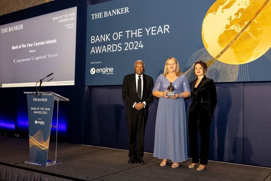 Three people stand on stage at the Bank of the Year Awards 2024, holding a trophy. A banner reads "Bank of the Year Cayman Islands - Cayman Capital Trust Bank.